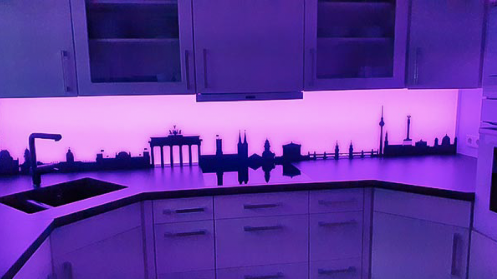 Küchenarbeitsplatte mit Berliner Skyline-Silhouette und violetter LED-Beleuchtung unter weißen Hängeschränken.