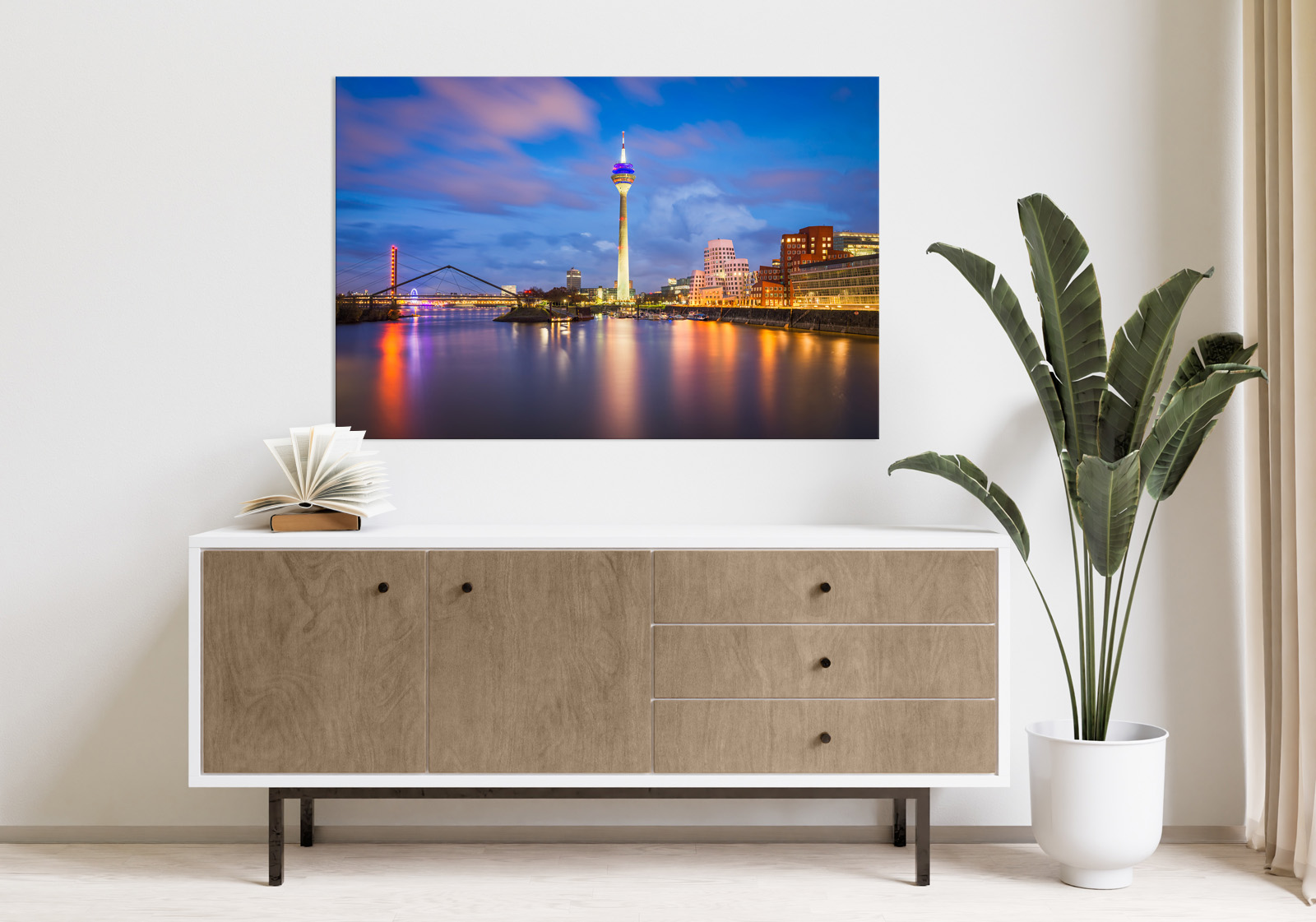 Milieubild mit hellem Holzsideboard mit Pflanze und darüber ein Wandbild mit folgendem Motiv: Düsseldorf-Skyline bei Nacht mit beleuchtetem Rheinturm, Brücke über den Rhein und reflektierenden Lichtern im Wasser.