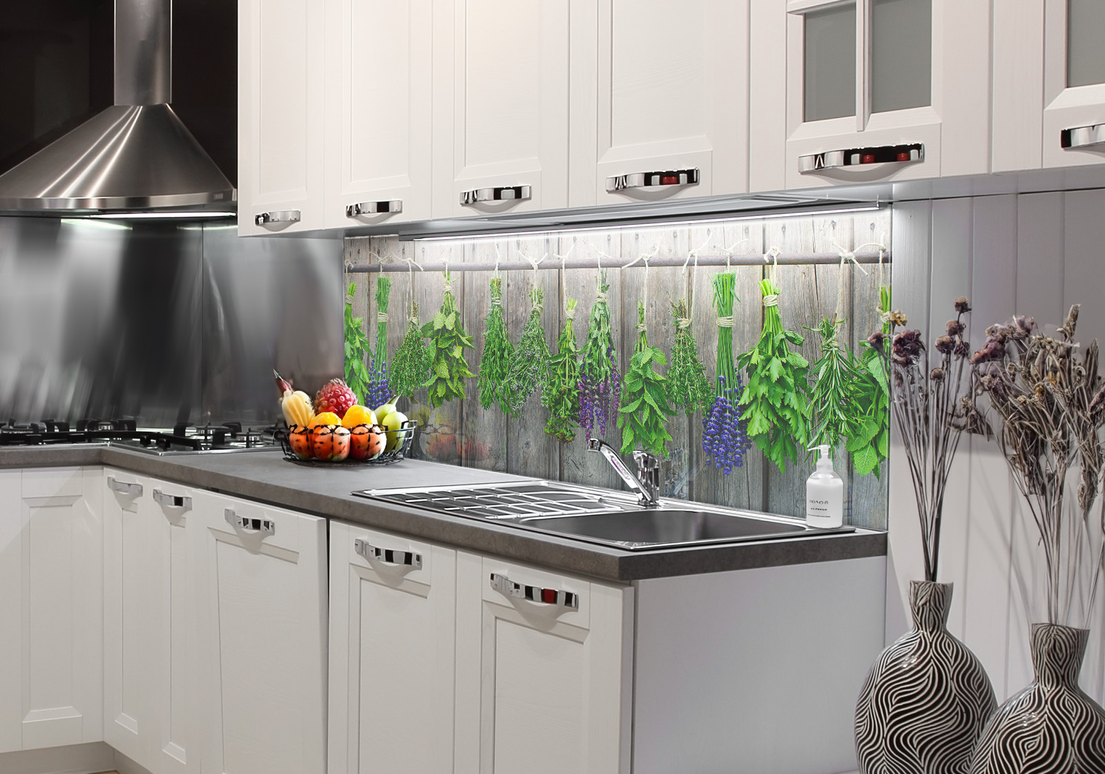 RW-K-P_Gewürze-03.jpg Weiße Küche mit  Edelstahl-Dunstabzugshaube, Spüle, Obstschale und zwei Vasen mit Dekozweigen. Es ist eine Küchenrückwand mit folgendem Motiv verbaut: Bündel von frischen Kräutern und lila Blüten hängen an einer Stange vor einer grauen Holzwand.