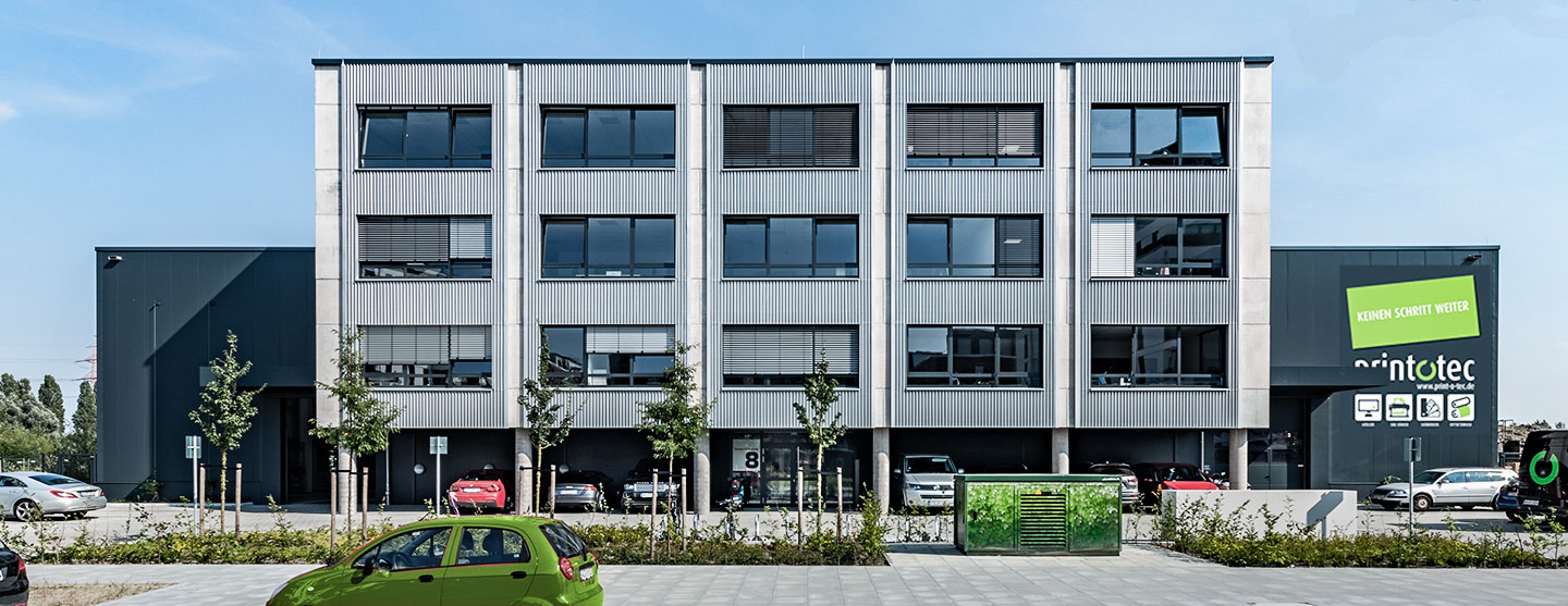 Außenansicht des Print-o-tec-Gebäudes mit grauer Fassade, Fensterfront und Parkplatz. Grüner Firmenname auf hellem Schild.