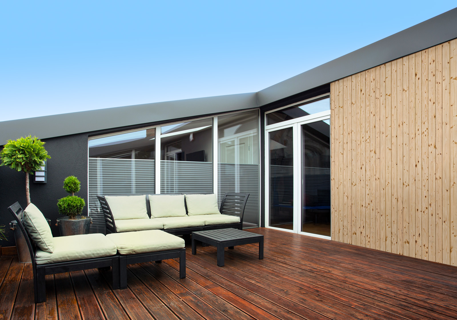 Milieubild mit Terrasse mit Holzdeck, Sitzgruppe mit hellen Polstern, großen Fenstern, grünen Pflanzen in Töpfen  und einer Wandverkleidung mit folgendem Motiv: Helle Holzplanken mit sichtbarer Maserung und Astlöchern, vertikal angeordnet.