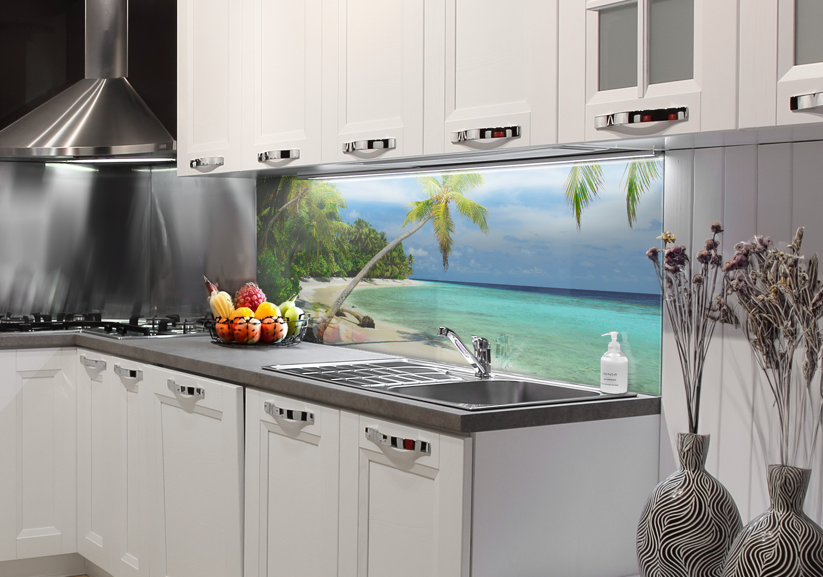 RW-K-P_Strand-02.jpg Weiße Küche mit Edelstahl-Dunstabzugshaube, Spüle, Obstschale und zwei Vasen mit Dekozweigen. Es ist eine Küchenrückwand mit folgendem Motiv verbaut: Tropischer Strand mit Palmen, weißem Sand und türkisfarbenem Wasser, sonniger Himmel mit Wolken.