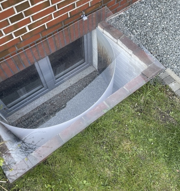 Blick von oben Lichtschacht mit Kellerfenster, abgedeckt durch eine gebogene transparente Kunststoffplatte, um Laub und Schmutz fernzuhalten.