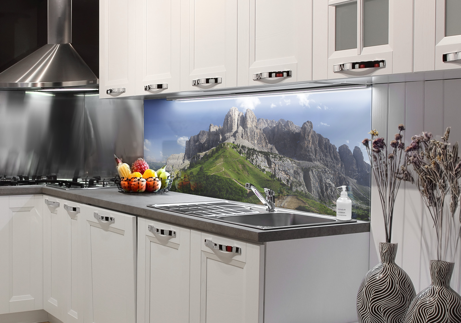 RW-K-P_Berge-01.jpg Weiße Küche mit  Edelstahl-Dunstabzugshaube, Spüle, Obstschale und zwei Vasen mit Dekozweigen. Es ist eine Küchenrückwand mit folgendem Motiv verbaut: Panorama der Dolomiten mit grünen Hängen und felsigen Gipfeln unter blauem Himmel.
