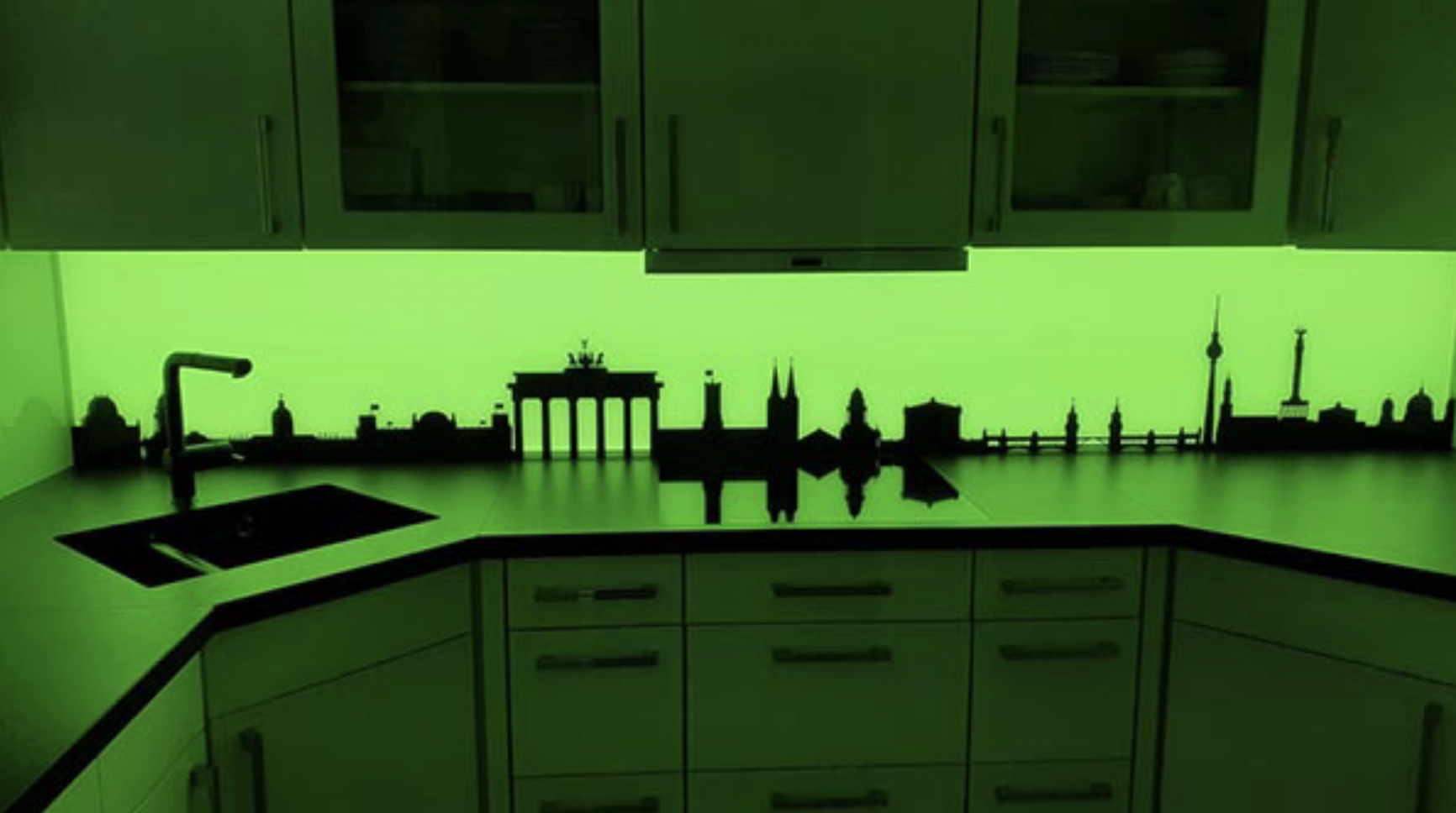 Küchenarbeitsplatte mit Berliner Skyline-Silhouette und grüner LED-Beleuchtung unter weißen Hängeschränken.