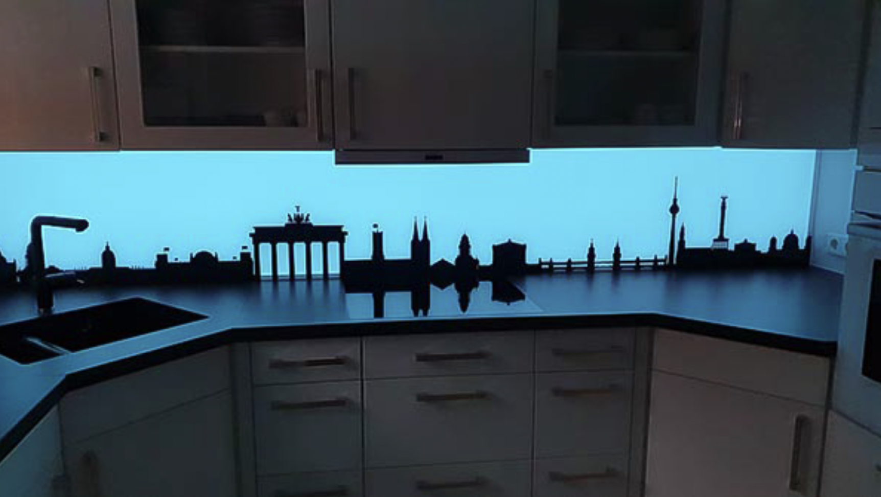 Küchenarbeitsplatte mit Berliner Skyline-Silhouette und blauer LED-Beleuchtung unter weißen Hängeschränken.