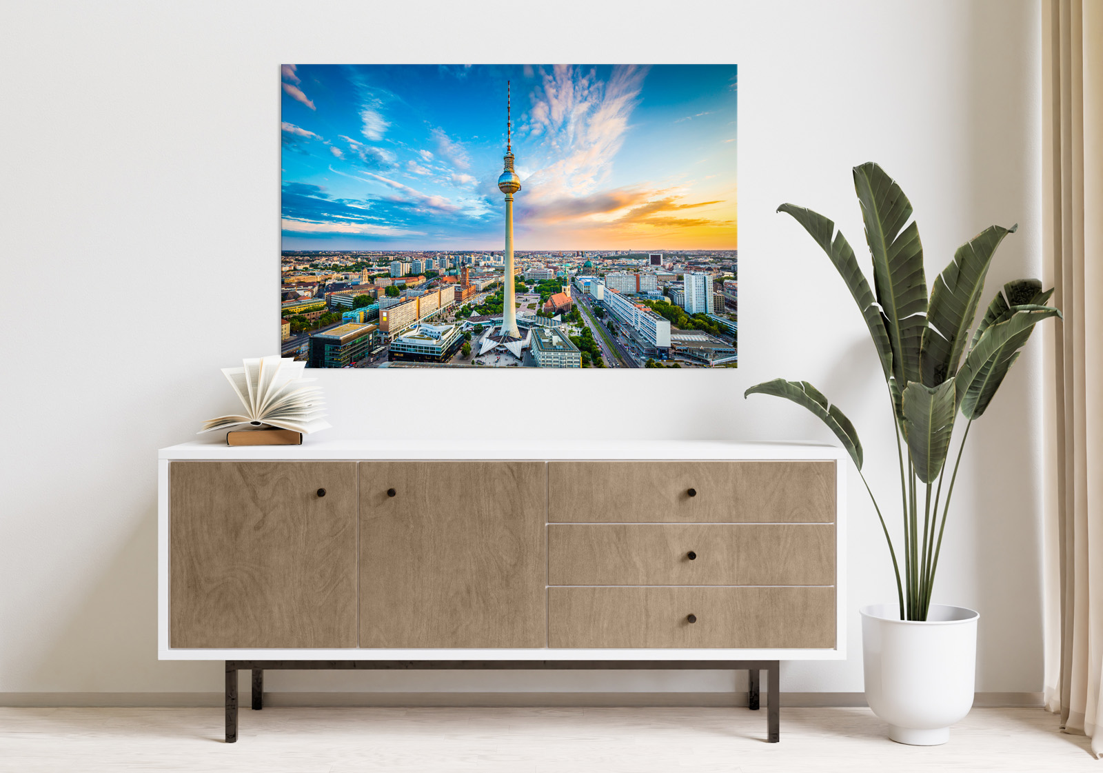 Milieubild mit hellem Holzsideboard mit Pflanze und darüber ein Wandbild mit folgendem Motiv: Luftaufnahme von Berlin mit Fernsehturm im Zentrum, Stadtbild bei Sonnenuntergang und blauem Himmel mit Wolken.