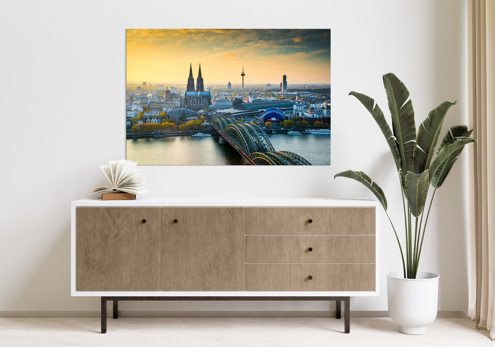 Milieubild mit hellem Holzsideboard mit Pflanze und darüber ein Wandbild mit folgendem Motiv: Kölner Dom und Hohenzollernbrücke bei Sonnenuntergang, Stadtbild mit Rhein und TV-Turm im Hintergrund.