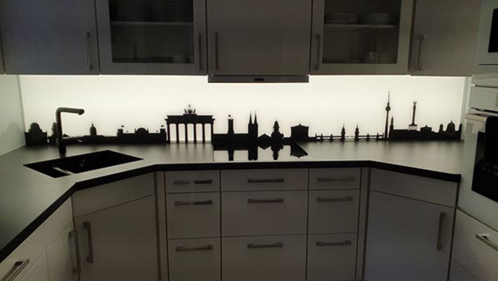 Küchenarbeitsplatte mit Berliner Skyline-Silhouette und weißer LED-Beleuchtung unter weißen Hängeschränken.