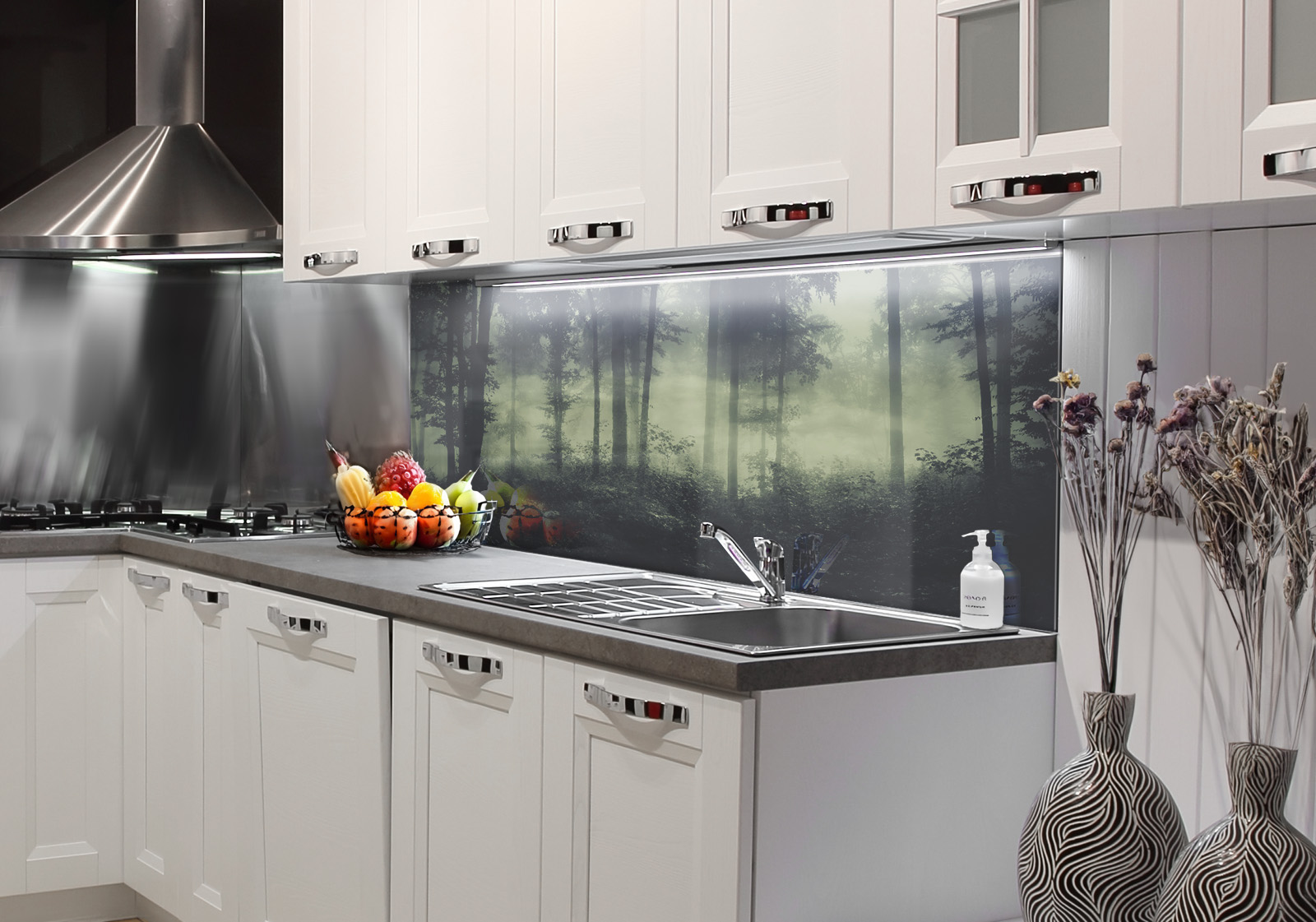 RW-K-P_Wald-02.jpg Weiße Küche mit Edelstahl-Dunstabzugshaube, Spüle, Obstschale und zwei Vasen mit Dekozweigen. Es ist eine Küchenrückwand mit folgendem Motiv verbaut: Dunkler Wald mit Nebel, hohe Bäume und Unterholz, mystische Atmosphäre.