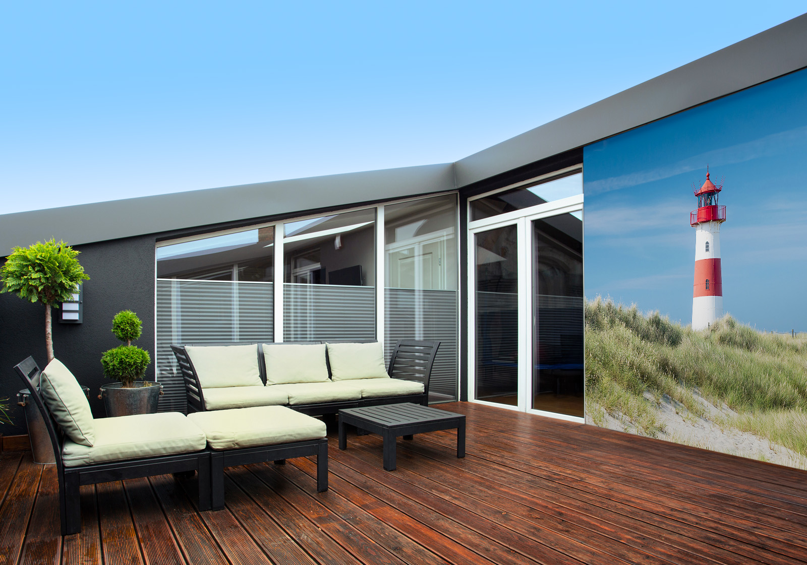 Milieubild mit Terrasse mit Holzdeck, Sitzgruppe mit hellen Polstern, großen Fenstern, grünen Pflanzen in Töpfen  und einer Wandverkleidung mit folgendem Motiv: Leuchtturm in rot und weiß vor blauem Himmel, im Vordergrund Dünen mit Dünengras und Zaun.