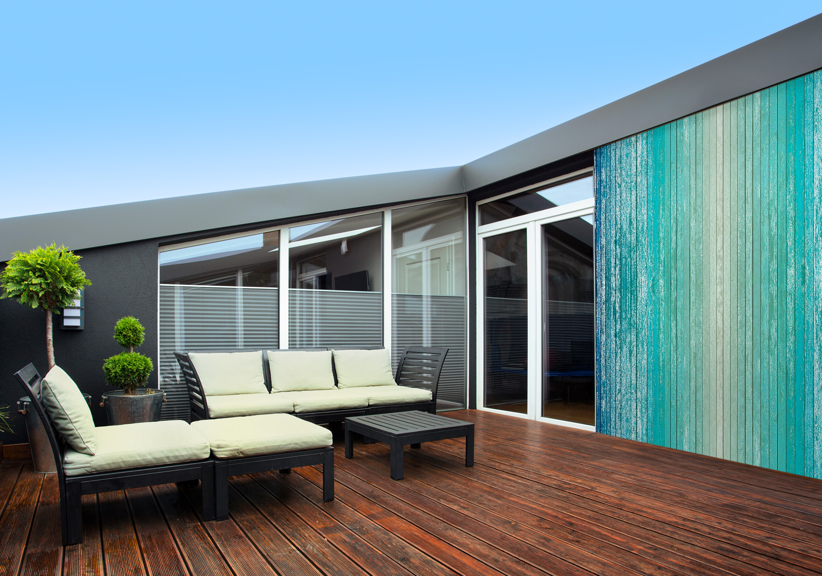 Milieubild mit Terrasse,Holzdeck, Sitzgruppe, hellen Polstern, großen Fenstern, grünen und einer Wandverkleidung mit Motiv: Holzplanken in Farbverlauf von Beige über Türkis bis Dunkelblau, mit sichtbarer Maserung und leichtem Vintage-Effekt.