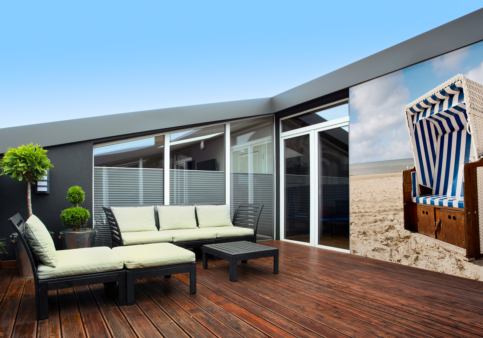 Milieubild mit Terrasse mit Holzdeck, Sitzgruppe mit hellen Polstern, großen Fenstern, grünen Pflanzen in Töpfen  und einer Wandverkleidung mit folgendem Motiv: Strandkorb mit blau-weißen Streifen auf Sandstrand vor ruhigem Meer und blauem Himmel.
