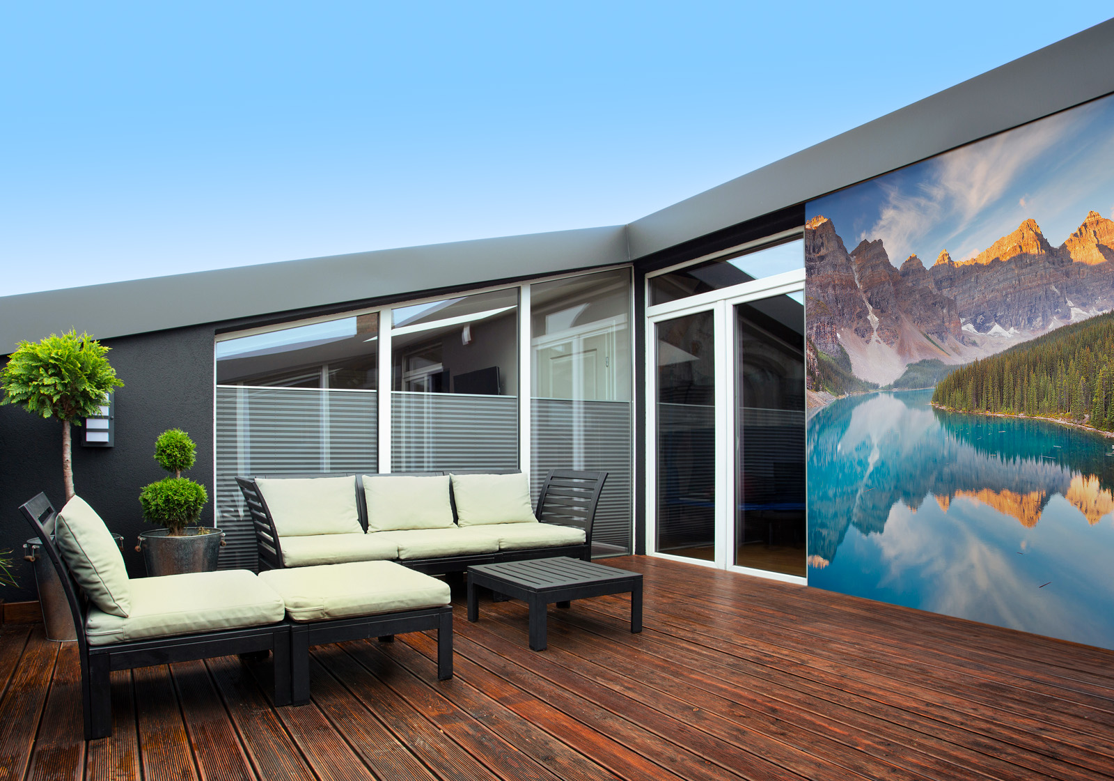 Milieubild mit Terrasse,Holzdeck, Sitzgruppe, hellen Polstern, großen Fenstern, grünen und einer Wandverkleidung mit Motiv: Spiegelung der schneebedeckten Berge im türkisfarbenen Wasser des Moraine Lake in Kanada, umgeben von dichten Nadelbäumen.