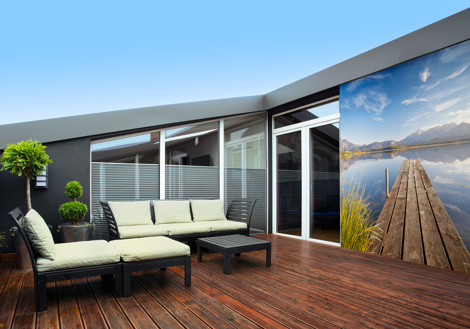 Milieubild mit Terrasse mit Holzdeck, Sitzgruppe mit hellen Polstern, großen Fenstern, grünen Pflanzen in Töpfen  und einer Wandverkleidung mit folgendem Motiv: Holzsteg am Badesee mit im Wasser spiegelndem Bergpanorama, seitlich hohe Wassergräser.