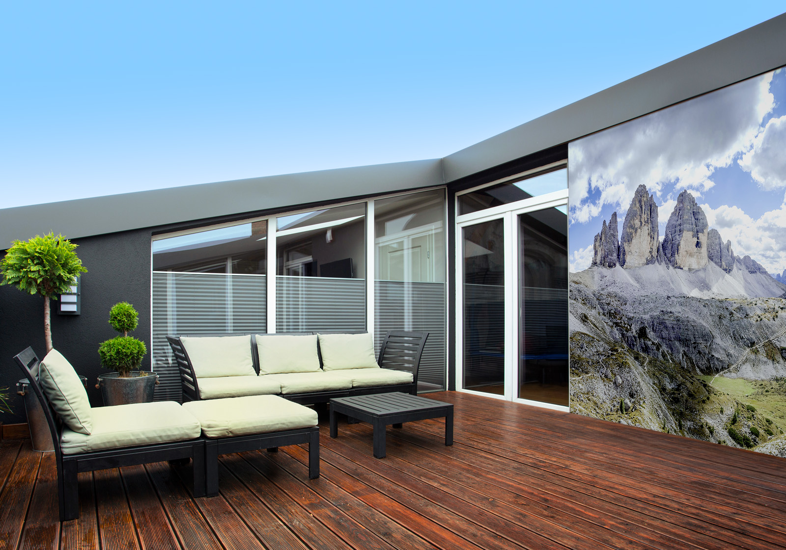Milieubild mit Terrasse,Holzdeck, Sitzgruppe, hellen Polstern, großen Fenstern, grünen und einer Wandverkleidung mit Motiv: Panorama der zerklüfteten Gipfel der Dolomiten unter einem Himmel mit flauschigen Wolken, mit einem kleinen See in einem Tal.