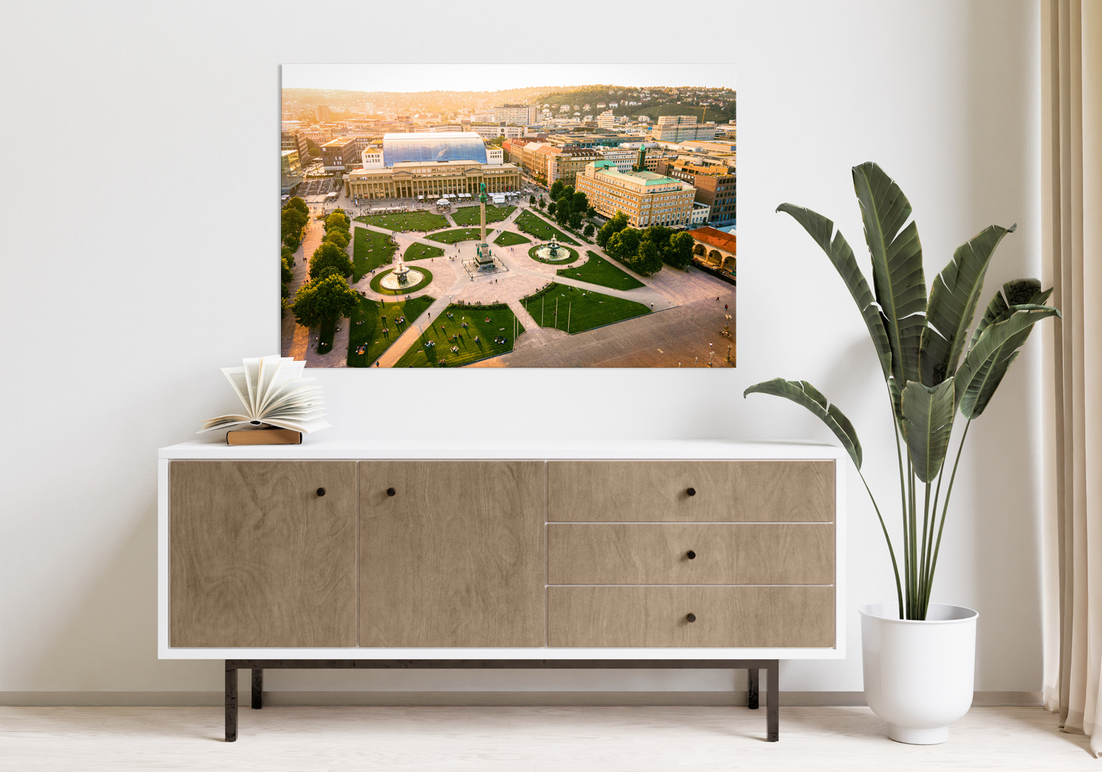 Milieubild mit hellem Holzsideboard mit Pflanze und darüber ein Wandbild mit folgendem Motiv: Luftaufnahme des Schlossplatzes Stuttgart mit Königsbau, Jubiläumssäule, Brunnen und Menschen, Sonnenuntergang.