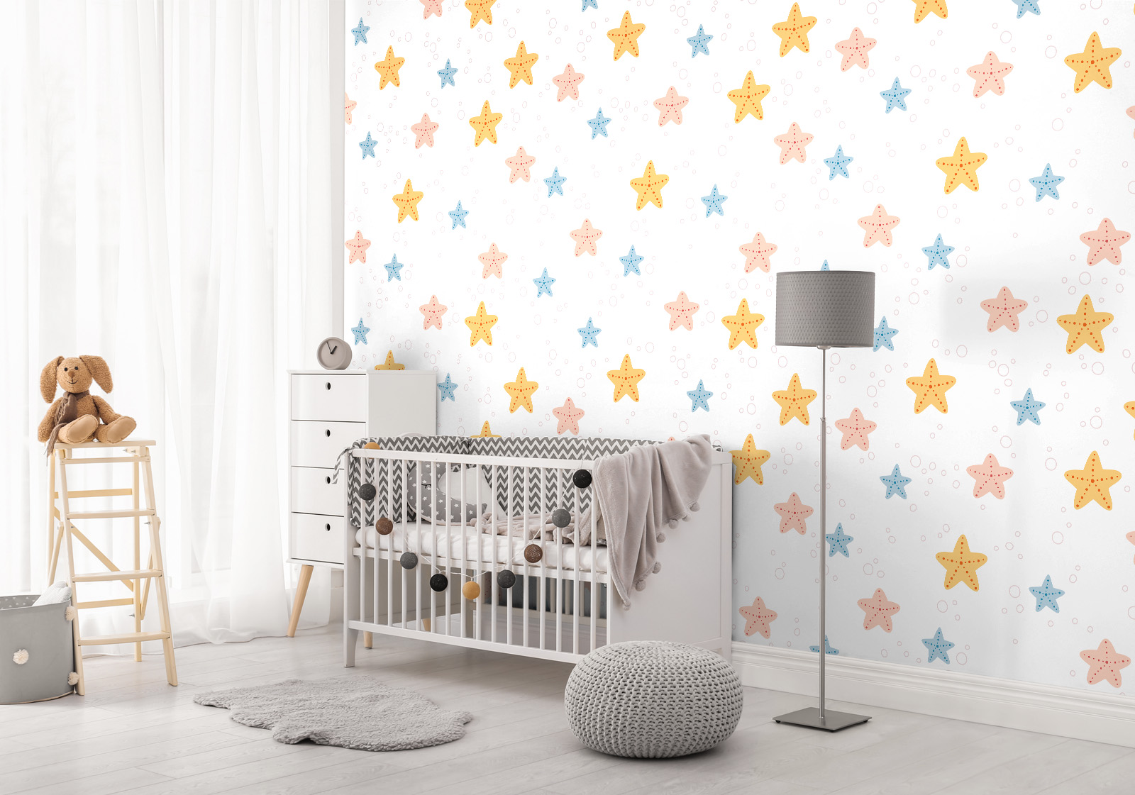 Milieubild mit  Kinderzimmer mit weißem Gitterbett, Stehlampe, Holzregal, grauem Teppich, Strickhocker und einer Wandverkleidung mit folgendem Motiv: Muster mit gelben, rosa und blauen Seesternen und kleinen Blasen auf weißem Hintergrund.