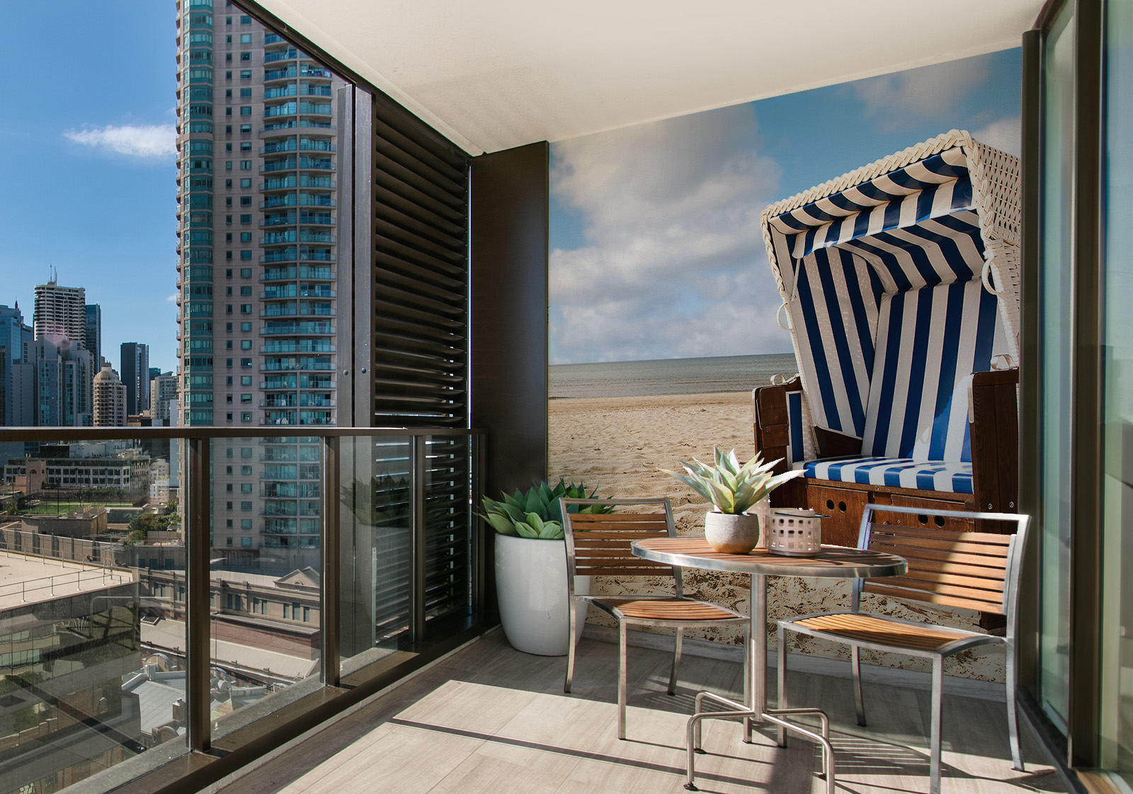 Milieubild mit Balkon mit Tisch und Stühlen, mit  Blick auf die Stadt und einer Wandverkleidung mit folgendem Motiv: Strandkorb mit blau-weißen Streifen auf Sandstrand vor ruhigem Meer und blauem Himmel.