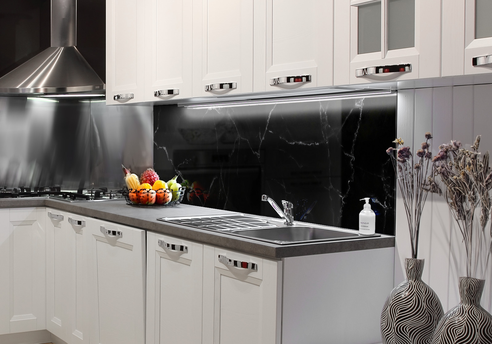 RW-K-P_Marmor-05.jpg Weiße Küche mit Edelstahl-Dunstabzugshaube, Spüle, Obstschale und zwei Vasen mit Dekozweigen. Es ist eine Küchenrückwand mit folgendem Motiv verbaut: Schwarzer Marmor mit weißen Adern, abstrakte Textur, dunkle und elegante Oberfläche.
