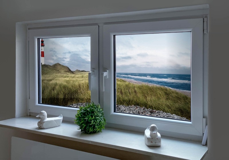 Hier klicken und entdecken Zwei Fenster mit einem Lichtschacht und Topfpflanze und zwei Entenfiguren auf der Fensterban. Im Lichtschacht ist ein Lichtschachtbild platziert mit folgendem Motiv: Panoramablick auf Strand mit Dünengras, Meer und Leuchtturm, bewölkter Himmel.