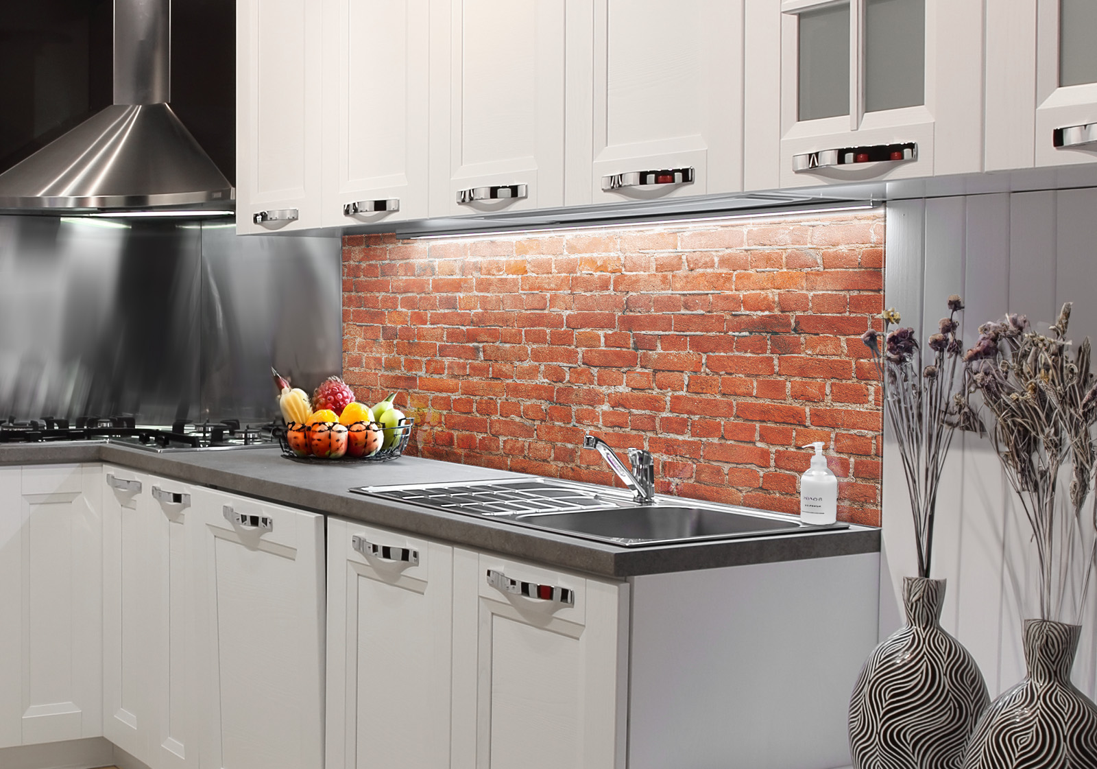 RW-K-P_Steine-02.jpg Weiße Küche mit Edelstahl-Dunstabzugshaube, Spüle, Obstschale und zwei Vasen mit Dekozweigen. Es ist eine Küchenrückwand mit folgendem Motiv verbaut: Rote Backsteinmauer mit grauen Fugen, regelmäßiges Muster, Textur.