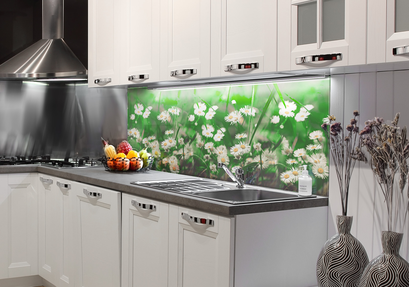 Weiße Küche mit  Edelstahl-Dunstabzugshaube, Spüle, Obstschale und zwei Vasen mit Dekozweigen. Es ist eine Küchenrückwand mit folgendem Motiv verbaut: Nahaufnahme von Gänseblümchen in einem grünen Feld mit weichem Fokus.