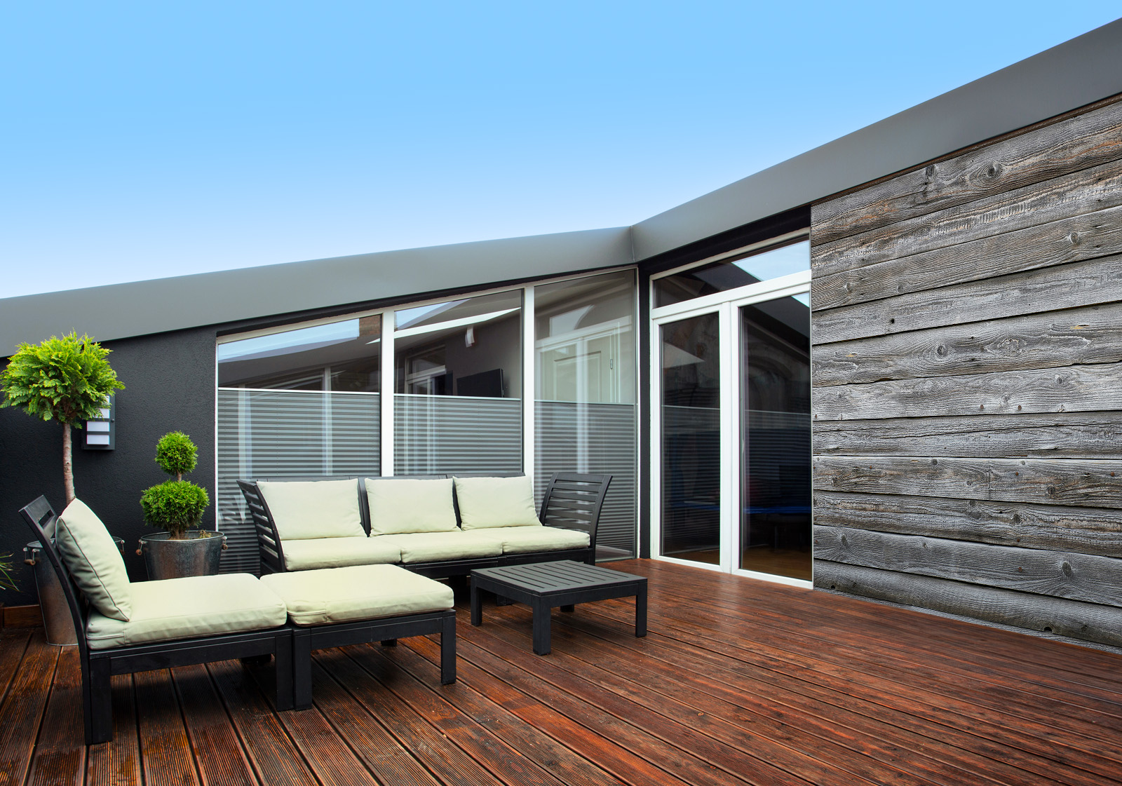 Milieubild mit Terrasse mit Holzdeck, Sitzgruppe mit hellen Polstern, großen Fenstern, grünen Pflanzen in Töpfen  und einer Wandverkleidung mit folgendem Motiv: Graue Holzplanken mit sichtbarer Maserung, symmetrisch angeordnet.