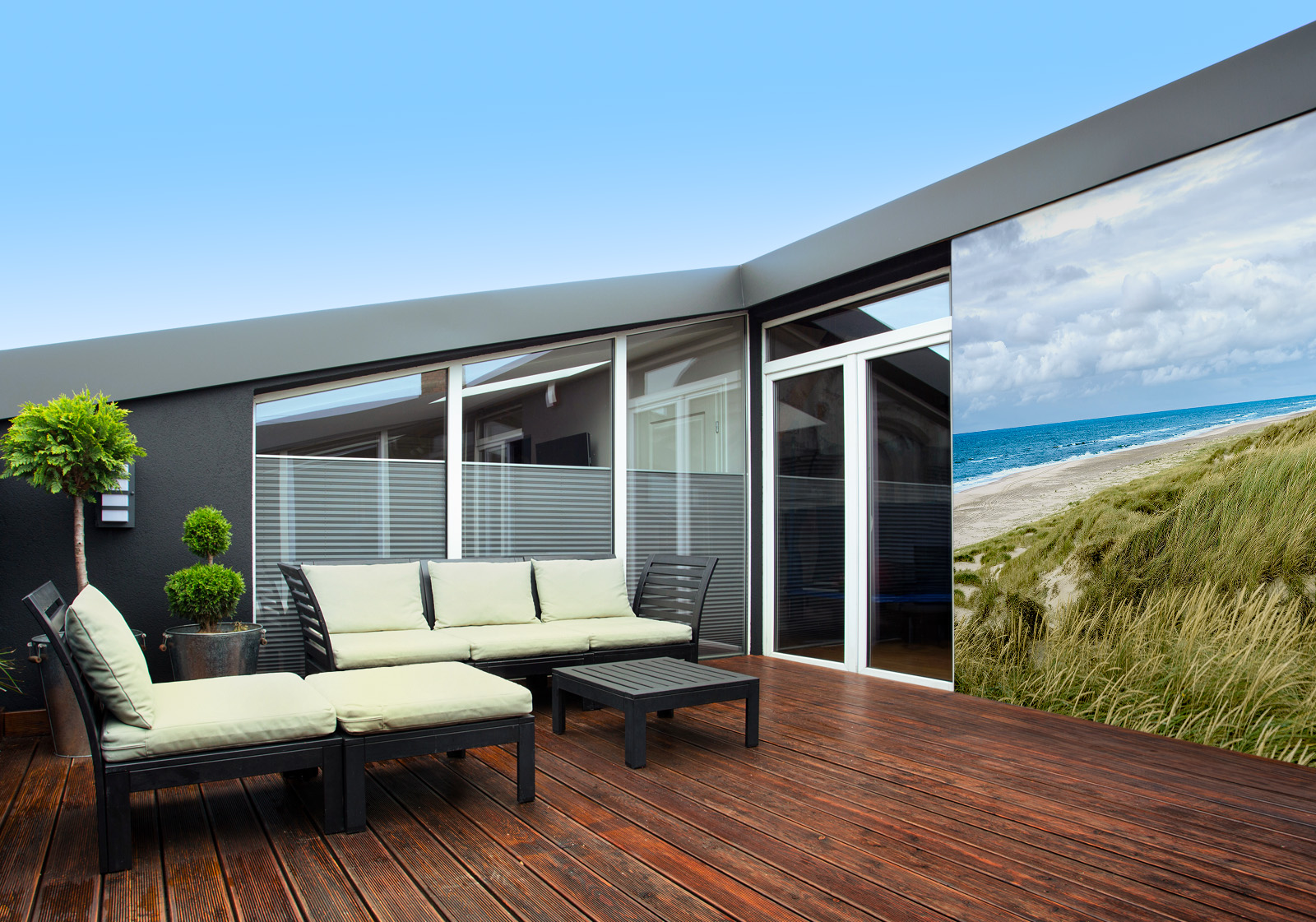 Milieubild mit Terrasse mit Holzdeck, Sitzgruppe mit hellen Polstern, großen Fenstern, grünen Pflanzen in Töpfen  und einer Wandverkleidung mit folgendem Motiv: Panoramablick auf Strand mit Dünengras, Meer und Leuchtturm, bewölkter Himmel.