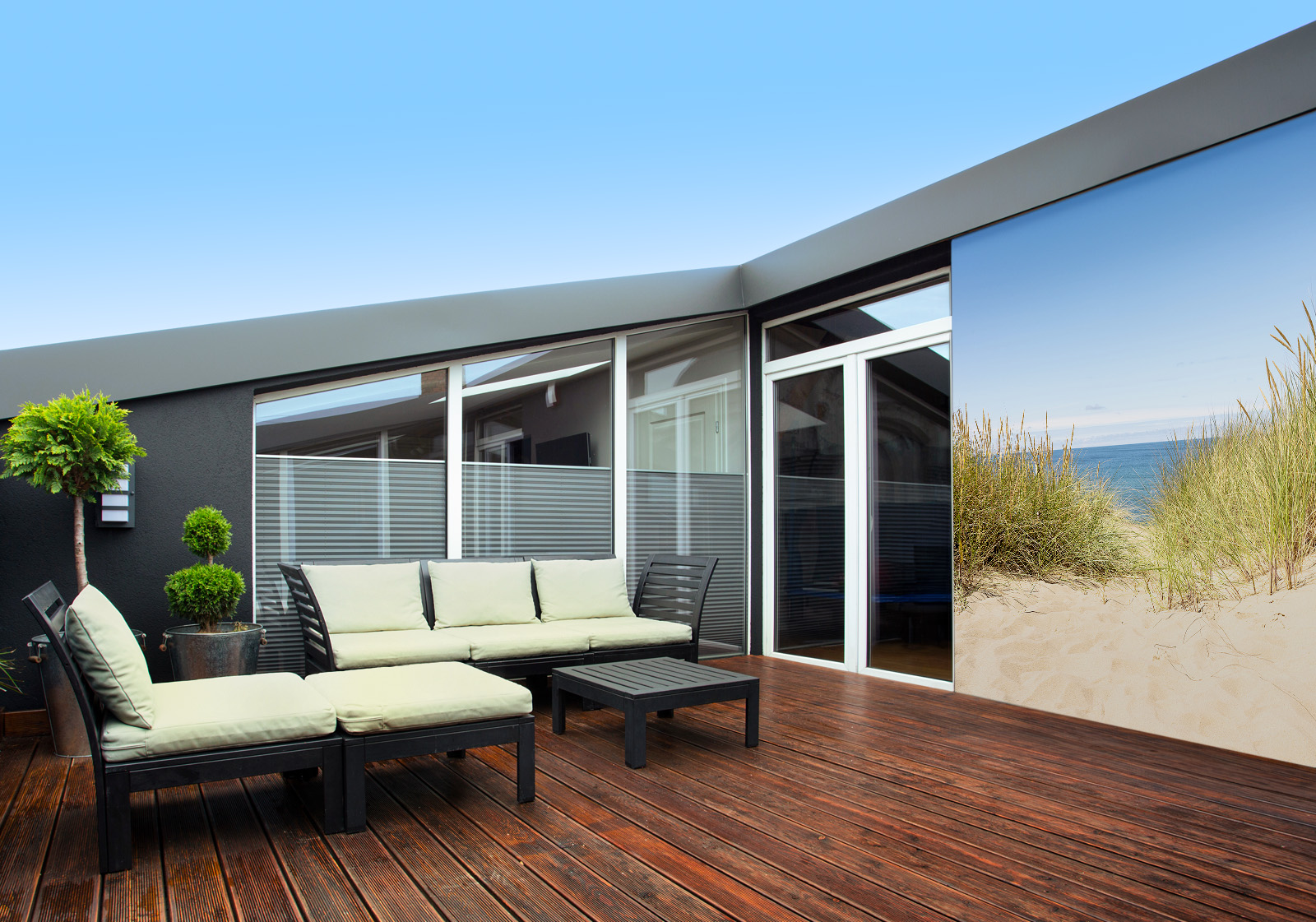 Milieubild mit Terrasse mit Holzdeck, Sitzgruppe mit hellen Polstern, großen Fenstern, grünen Pflanzen in Töpfen  und einer Wandverkleidung mit folgendem Motiv: Blick durch Dünengras auf den Sandstrand und das blaue Meer unter klarem Himmel.