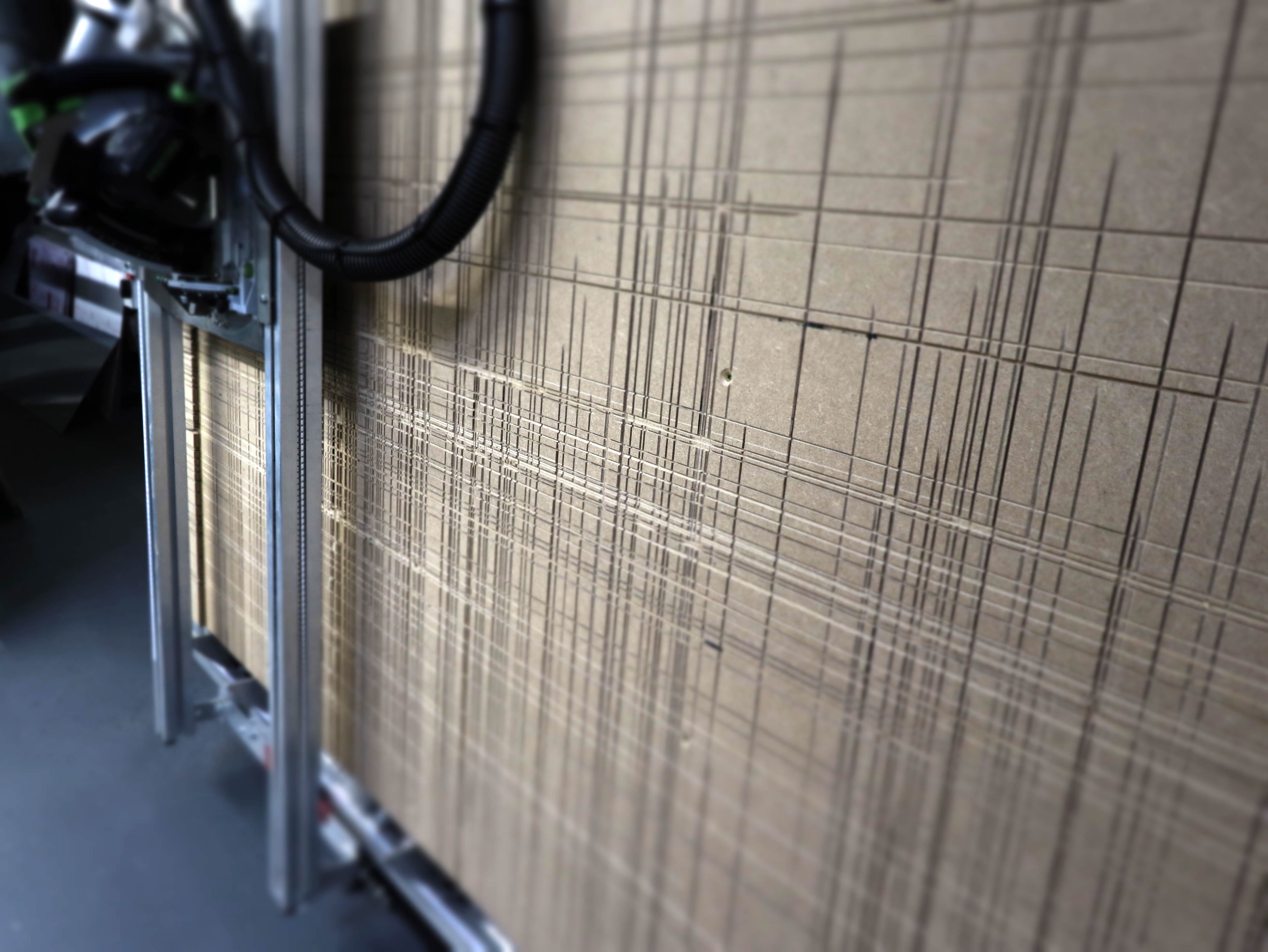Große, braune Platte mit feinen, parallelen Linien, die von einer Maschine geschnitten wurden. Im Vordergrund eine Metallschiene und ein schwarzer Schlauch.