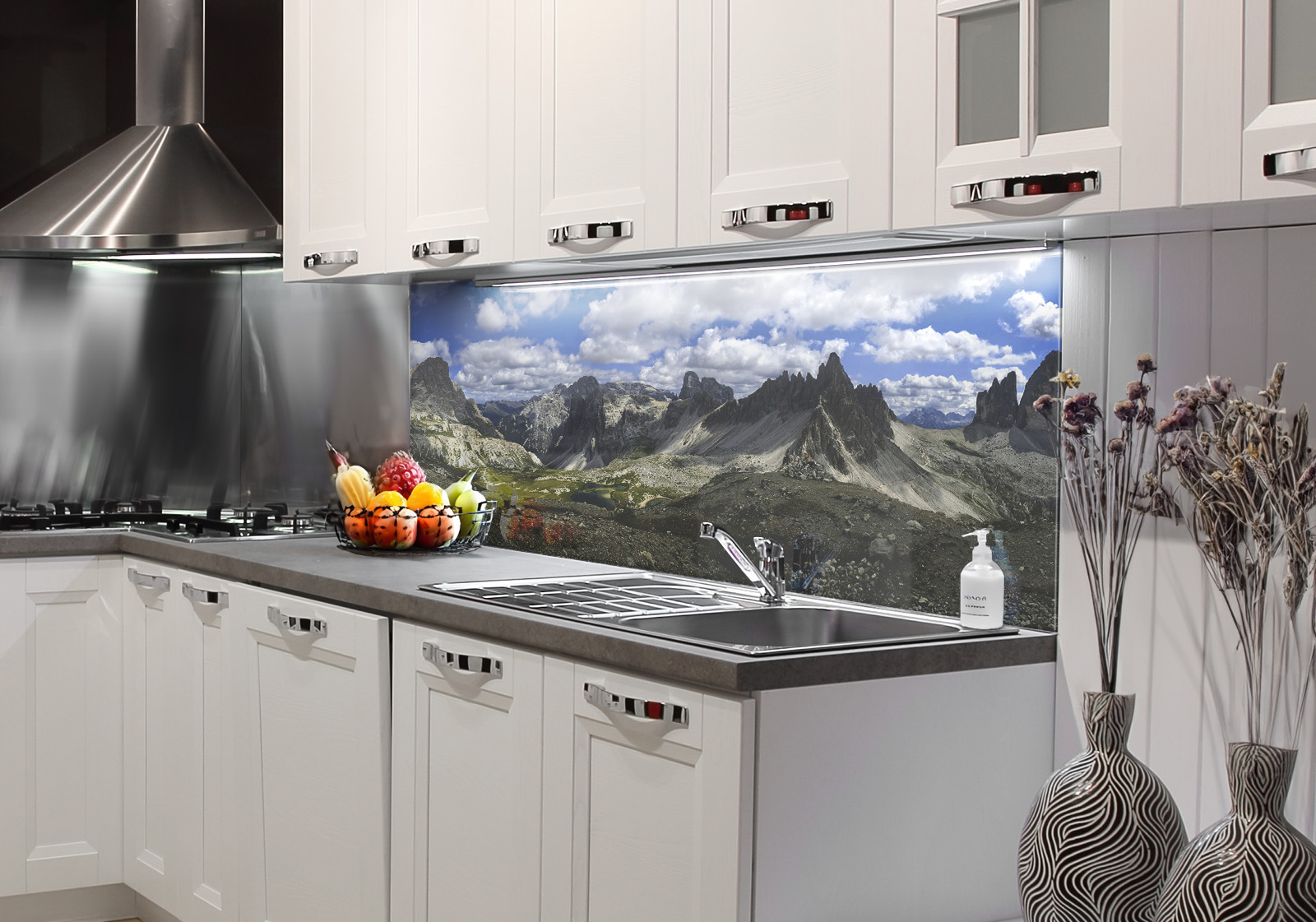 RW-K-P_Berge-04.jpg Weiße Küche mit  Edelstahl-Dunstabzugshaube, Spüle, Obstschale und zwei Vasen mit Dekozweigen und Küchenrückwand mit Motiv: Panorama der zerklüfteten Gipfel der Dolomiten unter einem Himmel mit flauschigen Wolken, mit einem kleinen See in einem Tal.