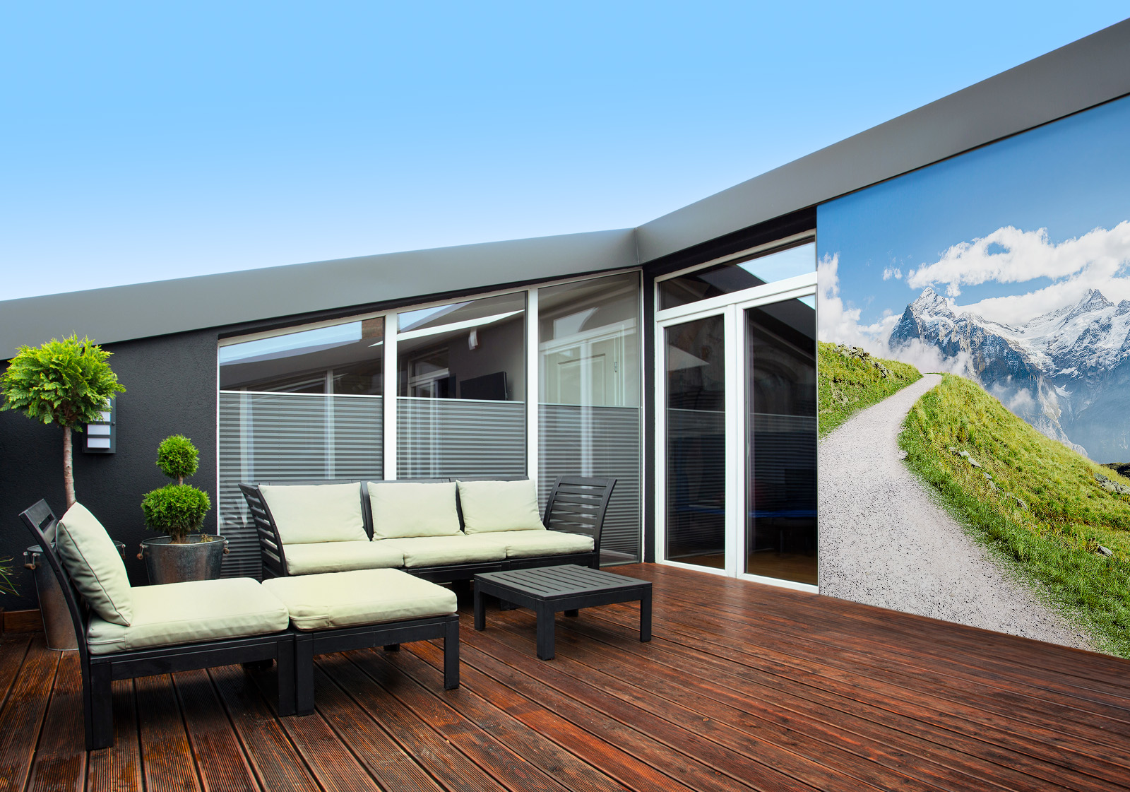 Milieubild mit Terrasse,Holzdeck, Sitzgruppe, hellen Polstern, großen Fenstern, grünen und einer Wandverkleidung mit Motiv: Wanderweg durch grüne Wiesen in den Schweizer Alpen, mit Blick auf schneebedeckte Berggipfel und blauen Himmel mit Wolken.