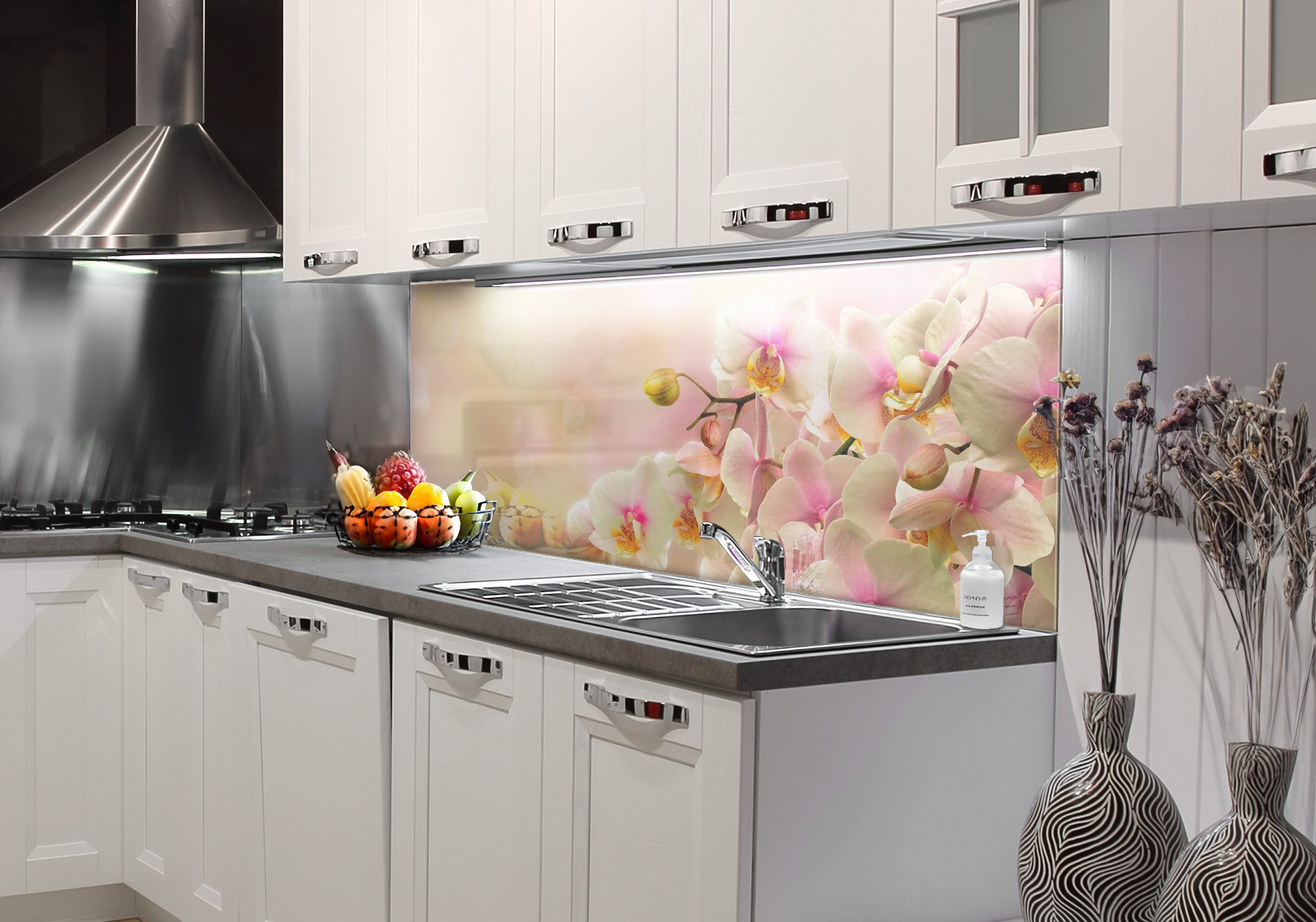 Weiße Küche mit  Edelstahl-Dunstabzugshaube, Spüle, Obstschale und zwei Vasen mit Dekozweigen. Es ist eine Küchenrückwand mit folgendem Motiv verbaut: Nahaufnahme von weißen Orchideenblüten mit rosa und gelben Akzenten, auf weichem, hellem Hintergrund.