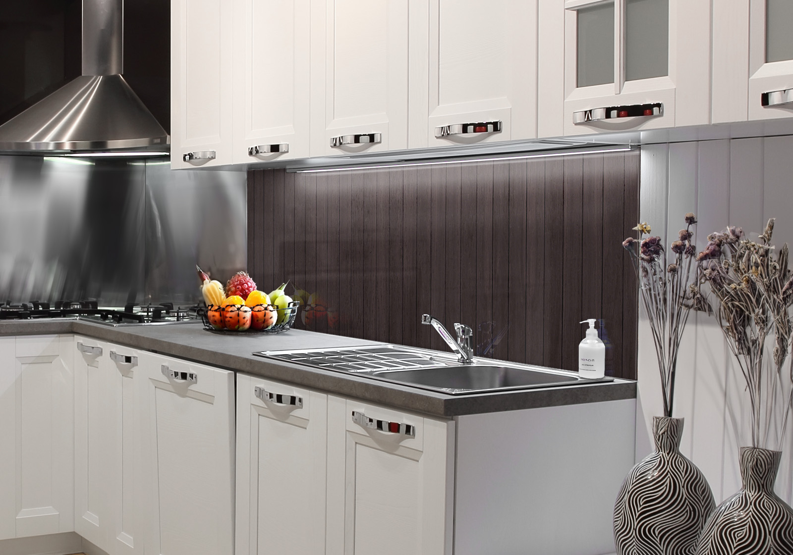 Weiße Küche mit Edelstahl-Dunstabzugshaube, Spüle, Obstschale und zwei Vasen mit Dekozweigen. Es ist eine Küchenrückwand mit folgendem Motiv verbaut: Dunkelbraune Holzpaneele mit feiner Maserung, vertikal angeordnet.