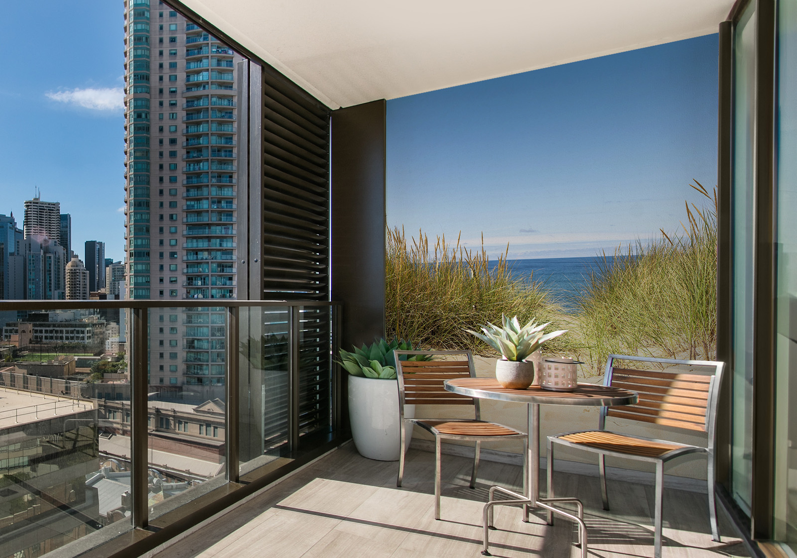 Milieubild mit Balkon mit Tisch und Stühlen, mit  Blick auf die Stadt und einer Wandverkleidung mit folgendem Motiv: Blick durch Dünengras auf den Sandstrand und das blaue Meer unter klarem Himmel.